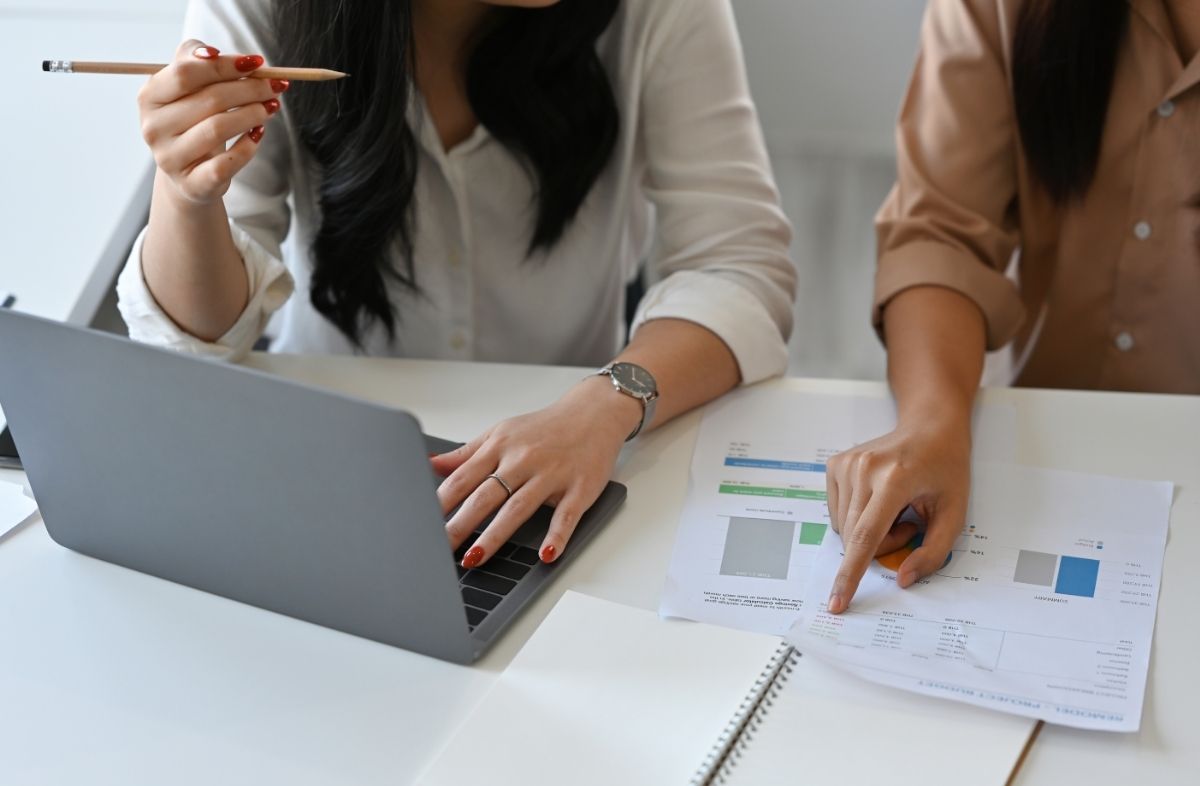 Dwie kobiety pracujące razem przy laptopie i analizujące wykresy na papierze